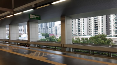 An elevated train station platform with a cityscape view in the background. Multiple high-rise residential buildings are visible, alongside lush green trees. The platform is modern with tiled floors and a sign indicating an exit direction in both English and Malay.