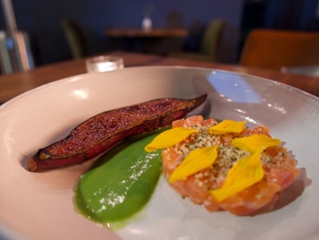 A beautifully plated dish with a piece of roasted eggplant on the left, a vibrant green puree in the middle, and a circular portion of salmon tartare garnished with yellow petals and spices on the right. The setting suggests a dimly lit restaurant atmosphere.