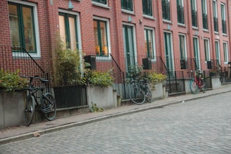 A row of red brick townhouses with white-framed windows and small front gardens bordered by wrought iron railings. Several bicycles are parked along the sidewalk, and small plants are visible in front of each house.