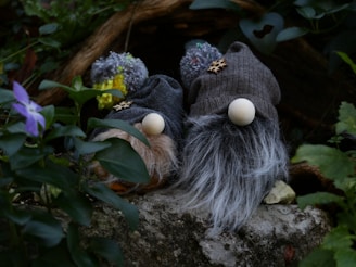 Two gnome-like figures with large hats and fluffy beards are nestled among greenery. Their hats are knitted with pom-poms on top, and the beards are made from thick, textured material. They rest on a stone surface surrounded by lush leaves and a purple flower in the foreground.
