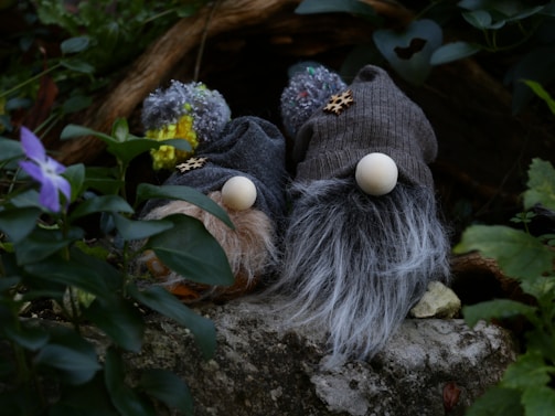 Two gnome-like figures with large hats and fluffy beards are nestled among greenery. Their hats are knitted with pom-poms on top, and the beards are made from thick, textured material. They rest on a stone surface surrounded by lush leaves and a purple flower in the foreground.