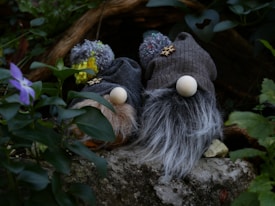 Two gnome-like figures with large hats and fluffy beards are nestled among greenery. Their hats are knitted with pom-poms on top, and the beards are made from thick, textured material. They rest on a stone surface surrounded by lush leaves and a purple flower in the foreground.