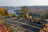 A scenic view of a forested landscape with a river meandering through the hills, framed by trees displaying vibrant autumn colors. A group of motorcyclists are parked along a road with a railing, taking in the picturesque view.