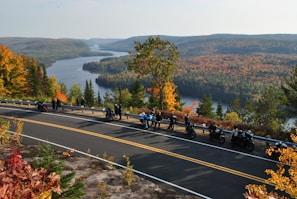 A scenic view of a forested landscape with a river meandering through the hills, framed by trees displaying vibrant autumn colors. A group of motorcyclists are parked along a road with a railing, taking in the picturesque view.