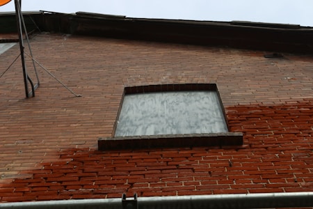 A brick wall with a boarded-up window and part of a satellite dish visible. The window is covered with a board that matches the reddish-brown tones of the brick. The bricks are laid in a running bond pattern, and some areas show signs of weathering and discoloration.