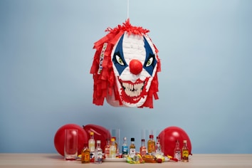 A clown-themed piñata with red hair and a sinister expression is suspended above a table. Below it, a collection of miniature liquor bottles, empty glassware, and two red balloons are arranged on the surface. The background is a plain light blue wall.