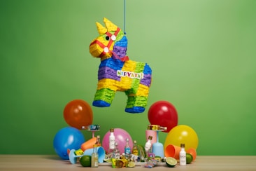 A colorful donkey-shaped piñata hangs against a vibrant green background. Below the piñata, there are several small bottles of alcohol, a mixture of festive balloons in blue, orange, red, and yellow, as well as assorted party items like candies and plastic cups.
