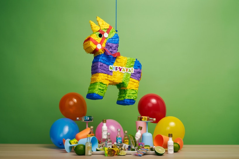 A colorful donkey-shaped pi&ntilde;ata hangs against a vibrant green background. Below the pi&ntilde;ata, there are several small bottles of alcohol, a mixture of festive balloons in blue, orange, red, and yellow, as well as assorted party items like candies and plastic cups.