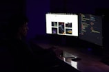 A person sits at a desk in a dimly lit room, facing two large computer monitors. One screen displays lines of colorful code, while the other shows a webpage with multiple images. The ambient lighting creates a focused and immersive atmosphere.