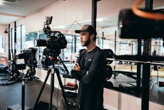 A person wearing a cap and casual clothing is standing indoors, near professional video recording equipment, possibly in a gym or fitness studio with exercise machines in the background.