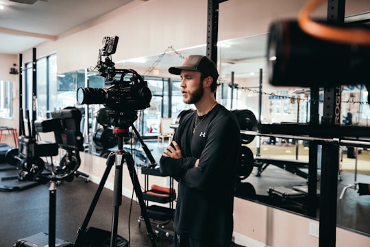 A person wearing a cap and casual clothing is standing indoors, near professional video recording equipment, possibly in a gym or fitness studio with exercise machines in the background.
