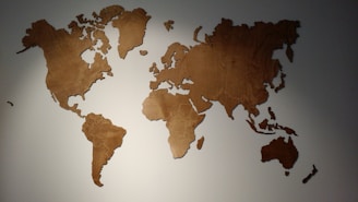 A wooden wall decoration in the shape of a world map with each continent cut out separately. The map is mounted on a light-colored wall and is illuminated from above, casting subtle shadows.