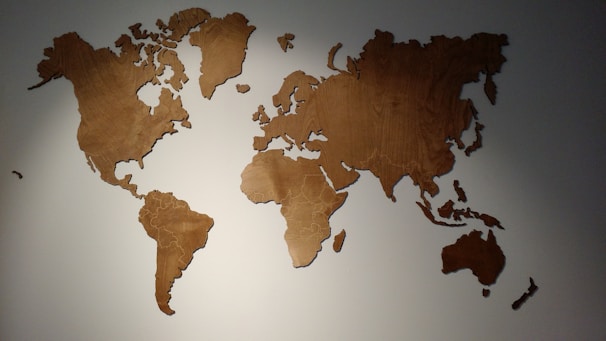 A wooden wall decoration in the shape of a world map with each continent cut out separately. The map is mounted on a light-colored wall and is illuminated from above, casting subtle shadows.