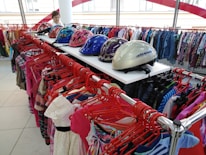 A clothing store display featuring a variety of colorful garments on red hangers. Above the clothing racks, there are several helmets with vibrant patterns, including pink with floral designs and a shiny silver one. A person in the background appears to be browsing the clothing.