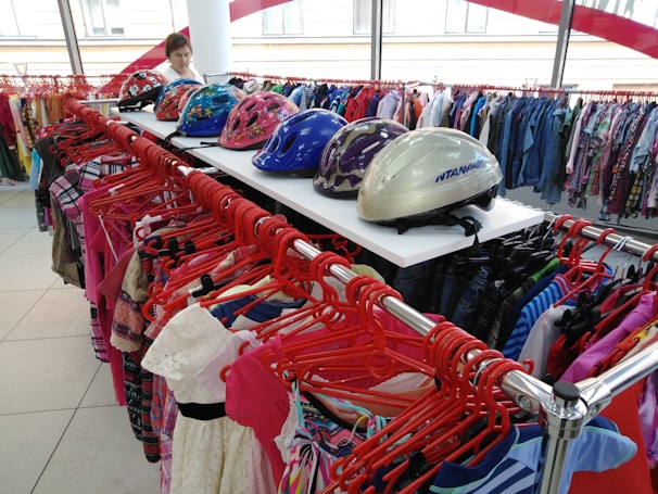 A clothing store display featuring a variety of colorful garments on red hangers. Above the clothing racks, there are several helmets with vibrant patterns, including pink with floral designs and a shiny silver one. A person in the background appears to be browsing the clothing.