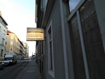 A street view with buildings lining both sides, featuring a prominent sign hanging from a building. The sign displays text in multiple languages, advertising a Russian-Armenian grocery. A white van and some cars are on the road, and the streets have a calm, urban atmosphere.