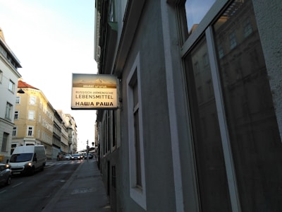 A street view with buildings lining both sides, featuring a prominent sign hanging from a building. The sign displays text in multiple languages, advertising a Russian-Armenian grocery. A white van and some cars are on the road, and the streets have a calm, urban atmosphere.