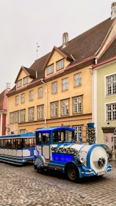 A festive tour train adorned with garlands of greenery and decorations is parked on a cobblestone street. The train is blue and white, and features a locomotive front with a cabin and two trailing cars. It is positioned in front of a series of historic, colorful European buildings with distinct architecture. The buildings are painted in shades of yellow, green, and beige with red and brown roofs.