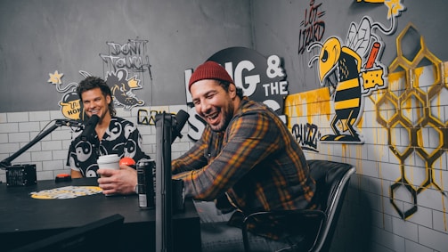 Two people are sitting in a studio with a lively and playful design. They are facing each other, with colorful graffiti featuring bees and honeycombs on the walls. The room has mounted microphones and they hold drinks, suggesting a podcast or radio setting.