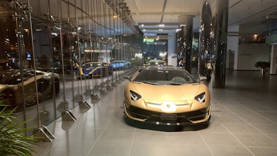 A luxurious sports car showroom with a prominent gold-colored car in the foreground. The showroom features reflective glass walls and a sleek, modern design. Several other high-end cars can be seen in the background, including a blue and yellow car. The showroom floor is clean and polished, and modern lighting fixtures enhance the upscale atmosphere of the space.