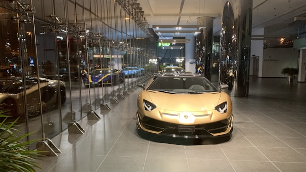 A luxurious sports car showroom with a prominent gold-colored car in the foreground. The showroom features reflective glass walls and a sleek, modern design. Several other high-end cars can be seen in the background, including a blue and yellow car. The showroom floor is clean and polished, and modern lighting fixtures enhance the upscale atmosphere of the space.