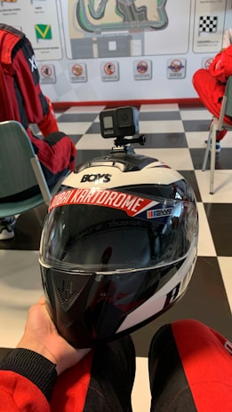 A racing helmet with a mounted action camera is held in hand. The helmet has the words 'Dubai Kartdrome' printed across the visor area. In the background, people dressed in red racing suits sit on chairs in a room with a checkered floor and a map on the wall.