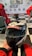 A racing helmet with a mounted action camera is held in hand. The helmet has the words 'Dubai Kartdrome' printed across the visor area. In the background, people dressed in red racing suits sit on chairs in a room with a checkered floor and a map on the wall.