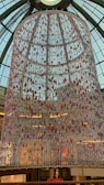 A large, cylindrical installation hangs from the ceiling of a building with a glass dome roof. The installation is composed of numerous strands adorned with red and white decorative elements. Light reflects off the hanging elements, creating a sparkling effect. Below, people can be seen walking inside a multi-level shopping mall. A sign for a restaurant is visible at the bottom.