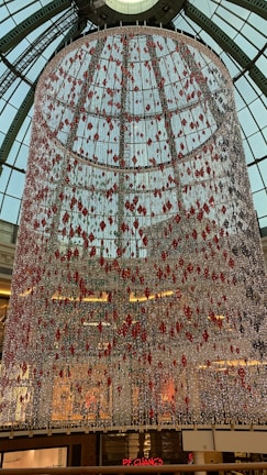 A large, cylindrical installation hangs from the ceiling of a building with a glass dome roof. The installation is composed of numerous strands adorned with red and white decorative elements. Light reflects off the hanging elements, creating a sparkling effect. Below, people can be seen walking inside a multi-level shopping mall. A sign for a restaurant is visible at the bottom.