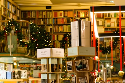 A cozy bookstore with shelves filled with a variety of books, adorned with festive garlands and small warmly-lit lights. Promotional signs for a 30% discount are visible. The atmosphere is enhanced by hanging decorations and organized displays, creating a warm and inviting environment.