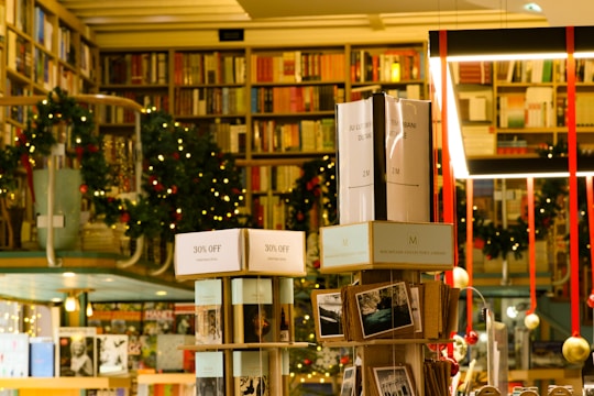 A cozy bookstore with shelves filled with a variety of books, adorned with festive garlands and small warmly-lit lights. Promotional signs for a 30% discount are visible. The atmosphere is enhanced by hanging decorations and organized displays, creating a warm and inviting environment.