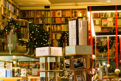 A cozy bookstore with shelves filled with a variety of books, adorned with festive garlands and small warmly-lit lights. Promotional signs for a 30% discount are visible. The atmosphere is enhanced by hanging decorations and organized displays, creating a warm and inviting environment.