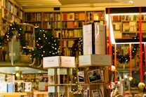 A cozy bookstore with shelves filled with a variety of books, adorned with festive garlands and small warmly-lit lights. Promotional signs for a 30% discount are visible. The atmosphere is enhanced by hanging decorations and organized displays, creating a warm and inviting environment.