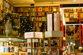A cozy bookstore with shelves filled with a variety of books, adorned with festive garlands and small warmly-lit lights. Promotional signs for a 30% discount are visible. The atmosphere is enhanced by hanging decorations and organized displays, creating a warm and inviting environment.