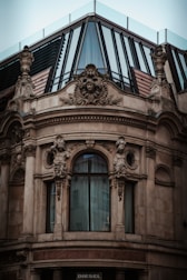 A richly adorned architectural facade features intricate sculptures and ornate carvings surrounding a large, arched window. The top section includes modern glass panels and metal framing, creating a contrast between classical and contemporary design elements.