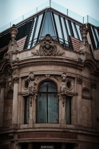 A richly adorned architectural facade features intricate sculptures and ornate carvings surrounding a large, arched window. The top section includes modern glass panels and metal framing, creating a contrast between classical and contemporary design elements.