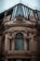 A richly adorned architectural facade features intricate sculptures and ornate carvings surrounding a large, arched window. The top section includes modern glass panels and metal framing, creating a contrast between classical and contemporary design elements.