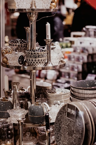 A display of intricately patterned ceramic bowls, teapots, and dishes organized on metallic shelves, all with a monochromatic color scheme featuring polka dots and stripes. Glass candlesticks and decorative items also appear on the shelves in an indoor store setting.