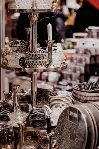 A display of intricately patterned ceramic bowls, teapots, and dishes organized on metallic shelves, all with a monochromatic color scheme featuring polka dots and stripes. Glass candlesticks and decorative items also appear on the shelves in an indoor store setting.