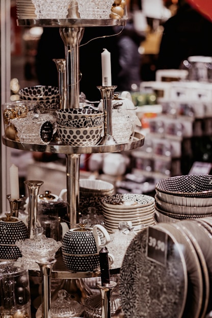 A display of intricately patterned ceramic bowls, teapots, and dishes organized on metallic shelves, all with a monochromatic color scheme featuring polka dots and stripes. Glass candlesticks and decorative items also appear on the shelves in an indoor store setting.