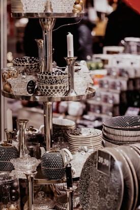 A display of intricately patterned ceramic bowls, teapots, and dishes organized on metallic shelves, all with a monochromatic color scheme featuring polka dots and stripes. Glass candlesticks and decorative items also appear on the shelves in an indoor store setting.