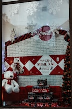 A festive store display featuring several large plush polar bears wearing Coca-Cola branded scarves. They are positioned against a red and white brick-patterned backdrop with a Coca-Cola logo. Decorative snowflakes hang above, and a Christmas tree adorned with baubles is partially visible. Numerous packs of Coca-Cola bottles are arranged in front of the bears.