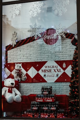 A festive store display featuring several large plush polar bears wearing Coca-Cola branded scarves. They are positioned against a red and white brick-patterned backdrop with a Coca-Cola logo. Decorative snowflakes hang above, and a Christmas tree adorned with baubles is partially visible. Numerous packs of Coca-Cola bottles are arranged in front of the bears.