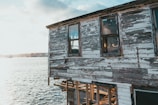 An old, weathered building with peeling paint stands next to a body of water. The structure's exterior is in poor condition, with visible wood rot and decay. A bright, sunlit sky is reflected on the water, creating a serene backdrop.