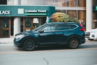 A green SUV is parked on a street with a large Christmas tree strapped to its roof. It is in front of a TD Canada Trust bank building. There's another car partially visible behind the SUV, and a person walking on the sidewalk.