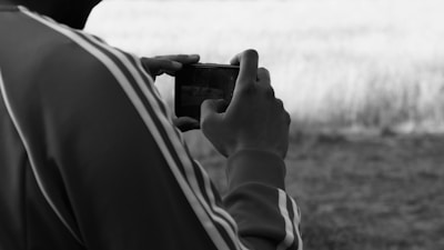 A person wearing a jacket with stripes on the sleeves holds a smartphone, pointing it at a field in front of them. The image is in black and white, focusing on the act of taking a photograph.