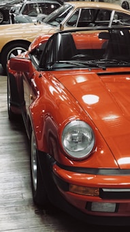 A vintage orange sports car is prominently displayed in the foreground, featuring classic rounded headlights and a sleek body design. In the background, several other classic cars are seen parked within a showroom environment, creating a nostalgic and elegant setting. The wooden floor adds to the vintage aesthetic.