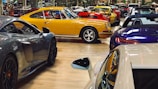 A collection of luxury sports cars are displayed in an indoor showroom with a polished wooden floor. The focus is on a vintage yellow car surrounded by modern vehicles of various colors, including red, blue, and gray. The setting appears affluent and neatly organized, highlighting the elegance and design of each car.