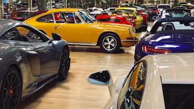A collection of luxury sports cars are displayed in an indoor showroom with a polished wooden floor. The focus is on a vintage yellow car surrounded by modern vehicles of various colors, including red, blue, and gray. The setting appears affluent and neatly organized, highlighting the elegance and design of each car.