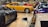 A collection of luxury sports cars are displayed in an indoor showroom with a polished wooden floor. The focus is on a vintage yellow car surrounded by modern vehicles of various colors, including red, blue, and gray. The setting appears affluent and neatly organized, highlighting the elegance and design of each car.